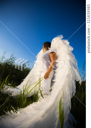 A woman with angel wings stands in a field, looking up at the sky. A magical scene that evokes hope and freedom. The sunlight bathes the angel in a bright glow, highlighting her purity and innocence. A woman with angel wings stands in a field, looking up at the sky. A magical scene that evokes hope and freedom. The sunlight bathes the angel in a bright glow, highlighting her purity and innocence. 121639365