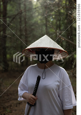 A man in a white shirt and a conical Asian hat holds a katana sword while standing in a forest. Rain falls on the trees in the background. A warrior with a katana stands in the forest in the rain. A man in a white shirt and a conical Asian hat holds a katana sword while standing in a forest. Rain falls on the trees in the background. A warrior with a katana stands in the forest in the rain. 121639366