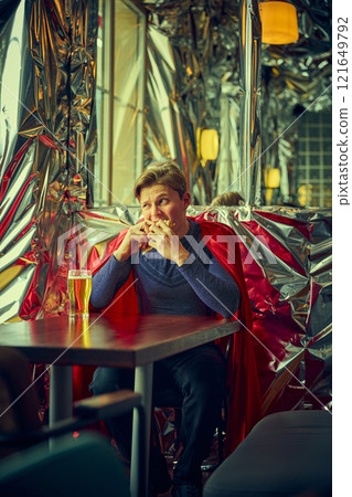 Man dressed as superhero pouring beer into glass, sitting in modern cafe with reflective walls, merging routine life with whimsical fantasy in vibrant surroundings. Man dressed as superhero pouring beer into glass, sitting in modern cafe with reflective walls, merging routine life with whimsical fantasy in vibrant surroundings. 121649792