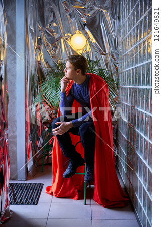 Superhero-dressed man sits by metallic wall, thoughtful gaze reflecting inner strength, futuristic vibes merging past inspiration and modern moments. 121649821