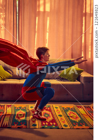 Mid-squat, cape flowing, superhero-themed man embodies modern balance between active lifestyle and grounded routine, surrounded by vibrant home decor. 121649823