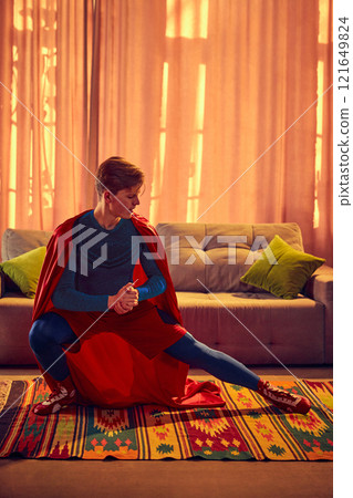 Superhero-clad man kneels low, doing exercises in living room at home in early morning, preparing for action or contemplation, 121649824
