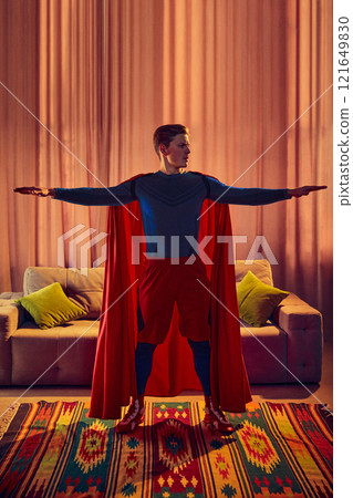 Man dressed as superhero stretches arms wide, training at home, to keep your body and spirit healthy, against warm tones of cozy living room. Man dressed as superhero stretches arms wide, training at home, to keep your body and spirit healthy, against warm tones of cozy living room. 121649830