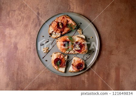 Sandwiches with thinly sliced salted salmon fillet, beetroot slices, walnuts, balsamic vinegar and fresh wheat bread on grey plate, fine dining. Brown background, top view. Colors of the year 2025. 121649834