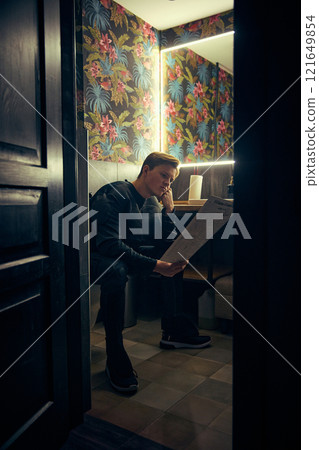 Man, dressed as superman, reading newspaper in dim bathroom, thoughtful break from day's tasks, finding peace in stillness. Man, dressed as superman, reading newspaper in dim bathroom, thoughtful break from day's tasks, finding peace in stillness. 121649854