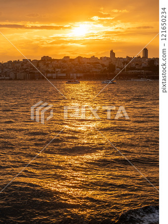 The sun sets over the Bosphorus, casting a golden glow on Istanbuls skyline while boats navigate the tranquil waters. The sun sets over the Bosphorus, casting a golden glow on Istanbuls skyline while boats navigate the tranquil waters. 121650234