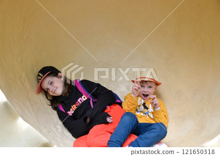 A boy and a girl happily sit on a slide in winter wearing helmets. They enjoy the leisure event, surrounded by a beautiful landscape, having fun and relaxing 121650318