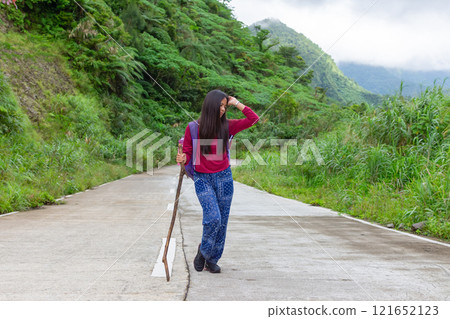 A tired young Asian woman walks along a mountain road, holding a stick in her hand, with a backpack, green hills in the background 121652123