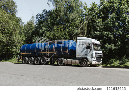 A truck with a white cabin and a blue tank parked on an asphalt area against the backdrop of green trees on a sunny day. A truck with a white cabin and a blue tank parked on an asphalt area against the backdrop of green trees on a sunny day. 121652613