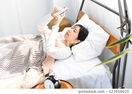 Smiling Asian woman looking at her smartphone in bed 121656020