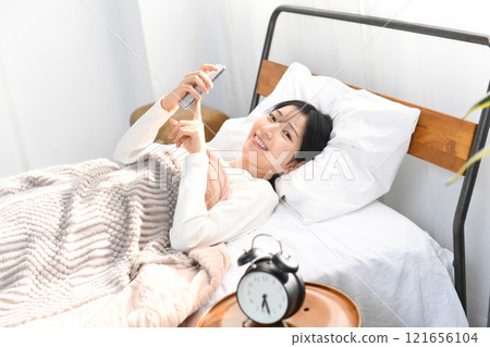 Smiling Asian woman holding smartphone in bed 121656104