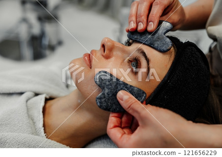 Woman receiving facial treatment with gua sha stones at spa during daytime relaxation session 121656229