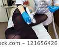 Close-up of a therapist beautician makes a LPG massage to a young woman in a dark transparent suit. Focus on the hands of a beautician with a maniple. 121656426