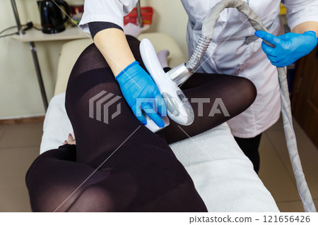 Close-up of a therapist beautician makes a LPG massage to a young woman in a dark transparent suit. Focus on the hands of a beautician with a maniple. 121656426