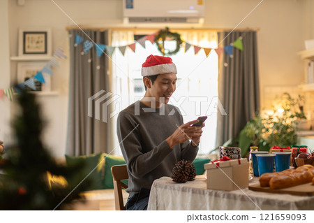 Man in Santa hat and casual sweater smiling while using smartphone at festively decorated table. Room is adorned with Christmas decorations, including garlands and colorful banners, as holiday treats 121659093