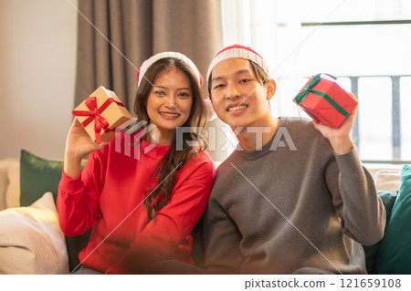 Cheerful Asian young couple lover both wearing red and white holiday hats, smiles while holding wrapped gifts. Woman dressed in red sweater and man in gray,They seated indoors on cozy couch 121659108