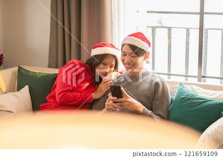 Asian young couple wearing festive Santa hats enjoys cozy moment on the couch, with the woman leaning affectionately on man shoulder while he smiles at smartphone. Holiday-themed setting adds warmth Asian young couple wearing festive Santa hats enjoys cozy moment on the couch, with the woman leaning affectionately on man shoulder while he smiles at smartphone. Holiday-themed setting adds warmth 121659109