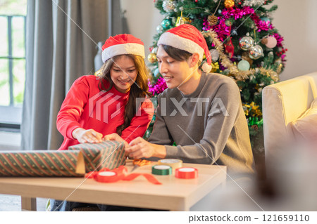 Joyful couple wearing Santa hats sits together near beautifully decorated Christmas tree, wrapping presents in festive wrapping paper. Holiday atmosphere warm and cheerful reflecting spirit of giving 121659110