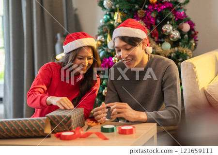 Joyful couple wearing Santa hats sits together near beautifully decorated Christmas tree, wrapping presents in festive wrapping paper. Holiday atmosphere warm and cheerful reflecting spirit of giving 121659111