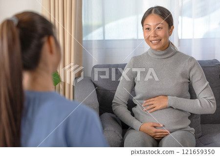 Asian pregnant woman is smiling during a prenatal consultation with a healthcare professional. Seated on sofa, gently touching her belly 121659350