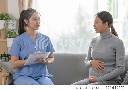 Asian pregnant woman is smiling during a prenatal consultation with a healthcare professional. Seated on sofa, gently touching her belly 121659351