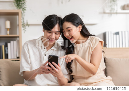 Asian young couple sit on couch or sofa looking at mobile phone smile and laughing together. Cheerful lover surfing internet and social media spending time in holiday at cozy home. Valentine Day 121659425