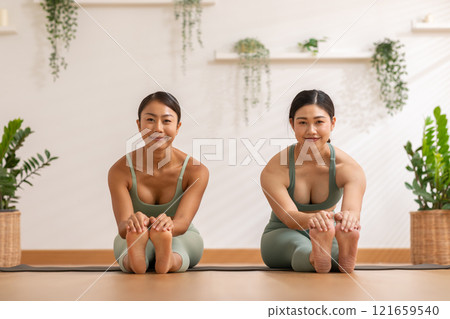 Two Asian women perform seated yoga stretch, bending gracefully to touch their feet showcasing flexibility and focus. They are dressed in matching green activewear with backdrop of natural plants Two Asian women perform seated yoga stretch, bending gracefully to touch their feet showcasing flexibility and focus. They are dressed in matching green activewear with backdrop of natural plants 121659540