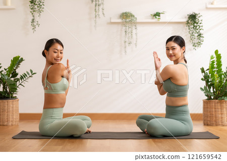 Two Asian women perform seated yoga stretch, bending gracefully to touch their feet showcasing flexibility and focus. They are dressed in matching green activewear with backdrop of natural plants 121659542