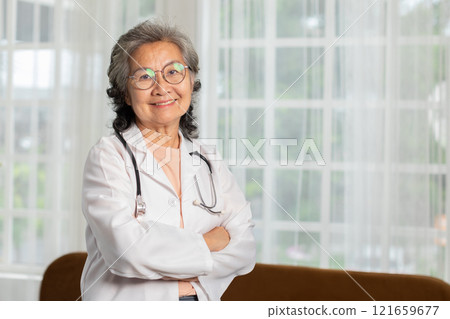 Smiling senior doctor with eyeglasses and stethoscope standing with arms crossed in front of large windows. 121659677