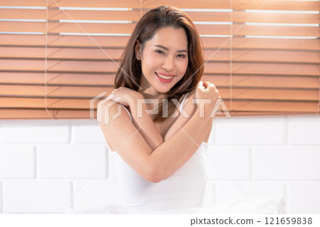 Cheerful Asian woman in white tank top, embracing herself with a warm and friendly smile. The background features wooden blinds, adding a cozy Cheerful Asian woman in white tank top, embracing herself with a warm and friendly smile. The background features wooden blinds, adding a cozy 121659838