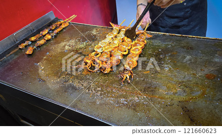 cook grills skewered squid over an open flame using a blowtorch, capturing the lively atmosphere of a bustling street food Thai market 121660623
