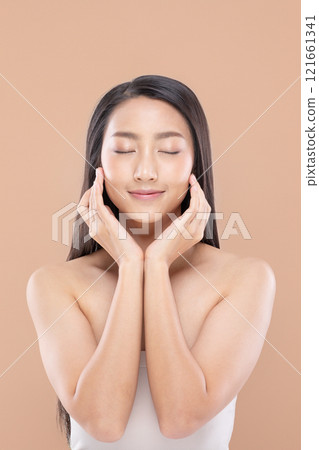 Serene and beautiful Asian young woman with flawless skin and long black hair. Gently touching her face with both hands, eyes closed, and smiling softly on soft peach color background Serene and beautiful Asian young woman with flawless skin and long black hair. Gently touching her face with both hands, eyes closed, and smiling softly on soft peach color background 121661341