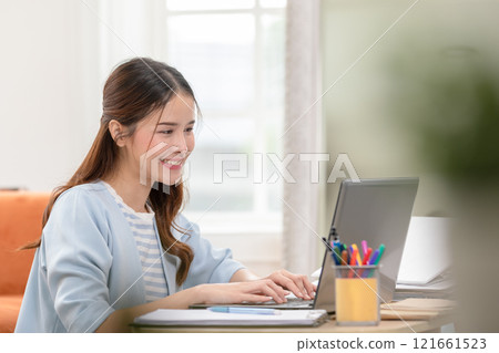 Asian Woman Smiling While Working on Laptop in Cozy Home Office Asian Woman Smiling While Working on Laptop in Cozy Home Office 121661523
