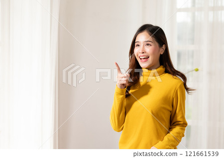 Smiling young woman in yellow sweater pointing upwards, expressing idea or inspiration, standing in brightly lit room. 121661539