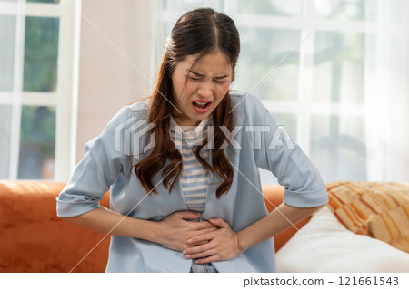 Woman experiencing stomach pain while sitting on the couch, expressing discomfort and holding her abdomen in a bright living room setting. Woman experiencing stomach pain while sitting on the couch, expressing discomfort and holding her abdomen in a bright living room setting. 121661543