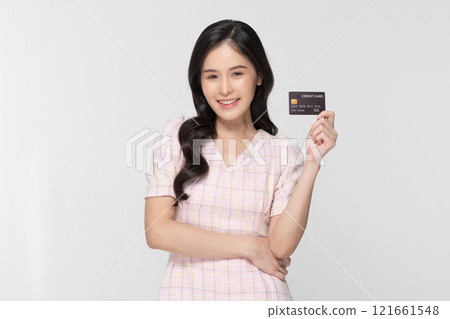 Cheerful Asian young woman confidently holds up credit card with bright smile against plain white background. Dressed in light stylish blouse exudes positivity and financial empowerment 121661548