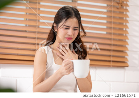 Asian woman in a white tank top holding a cup, showing discomfort and touching her throat, Health care concept Asian woman in a white tank top holding a cup, showing discomfort and touching her throat, Health care concept 121661571