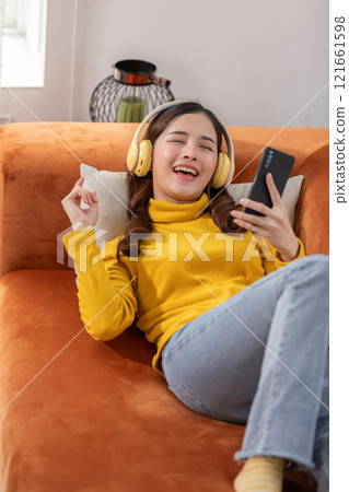 Joyful Asian young woman relaxing on an orange couch, wearing yellow headphones and a matching sweater, and holding a smartphone. She is smiling and enjoying the music Joyful Asian young woman relaxing on an orange couch, wearing yellow headphones and a matching sweater, and holding a smartphone. She is smiling and enjoying the music 121661598