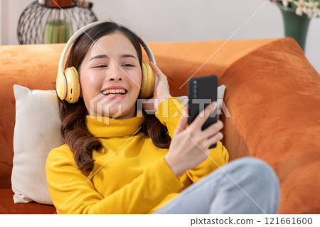 Joyful Asian young woman relaxing on an orange couch, wearing yellow headphones and a matching sweater, and holding a smartphone. She is smiling and enjoying the music 121661600