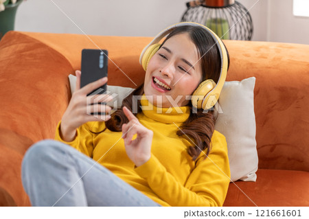 Joyful Asian young woman relaxing on an orange couch, wearing yellow headphones and a matching sweater, and holding a smartphone. She is smiling and enjoying the music Joyful Asian young woman relaxing on an orange couch, wearing yellow headphones and a matching sweater, and holding a smartphone. She is smiling and enjoying the music 121661601