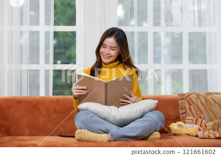 Content young Asian woman reading a book on a comfy sofa with a warm throw pillow. Content young Asian woman reading a book on a comfy sofa with a warm throw pillow. 121661602