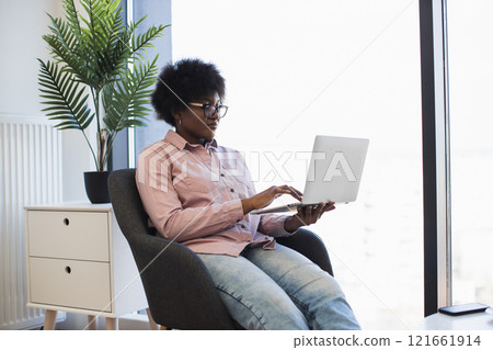 African American woman working on laptop in modern office setting. Professional business environment with natural light and contemporary decor. Focused and determined atmosphere. 121661914