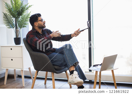 Indian man enjoying video call in modern home office. Wearing casual attire, sitting on chair, interacting with smartphone. Bright and airy atmosphere with large window and indoor plant. Indian man enjoying video call in modern home office. Wearing casual attire, sitting on chair, interacting with smartphone. Bright and airy atmosphere with large window and indoor plant. 121661922