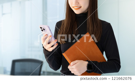 Smiling woman/businesswoman holding a smartphone and a binder 121662076