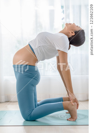 Pregnant woman practicing yoga in a bright room. She performs a camel pose on a mat, emphasizing health, well-being, and prenatal fitness. 121662109