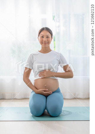 Pregnant woman practicing yoga in the living room, meditating in a comfortable pose, with natural light and peaceful ambiance. 121662110
