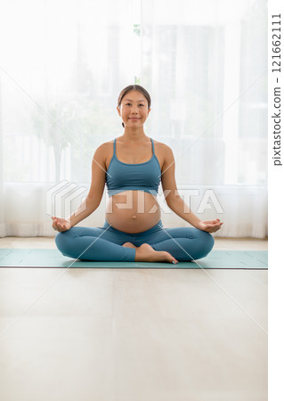 Pregnant woman practicing yoga indoors, meditating in lotus pose on mat. Prenatal exercise and healthy lifestyle concept. 121662111