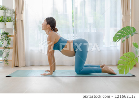 Pregnant woman practicing yoga indoors on a mat, wearing blue sportswear, in a light-filled room with plants and curtains. Pregnant woman practicing yoga indoors on a mat, wearing blue sportswear, in a light-filled room with plants and curtains. 121662112