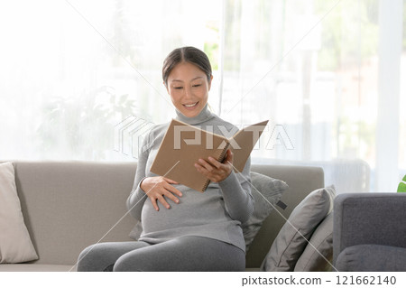 Asian pregnant woman is seated on a comfortable sofa, engrossed in reading a book. She gently touches her belly, Serene and reflective moments of pregnancy 121662140