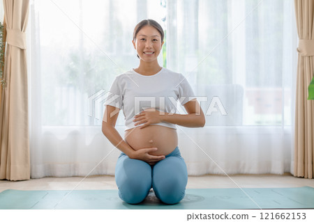 Pregnant woman practicing prenatal yoga at home, kneeling on a mat, holding her belly, and smiling peacefully during a bright morning. Pregnant woman practicing prenatal yoga at home, kneeling on a mat, holding her belly, and smiling peacefully during a bright morning. 121662153